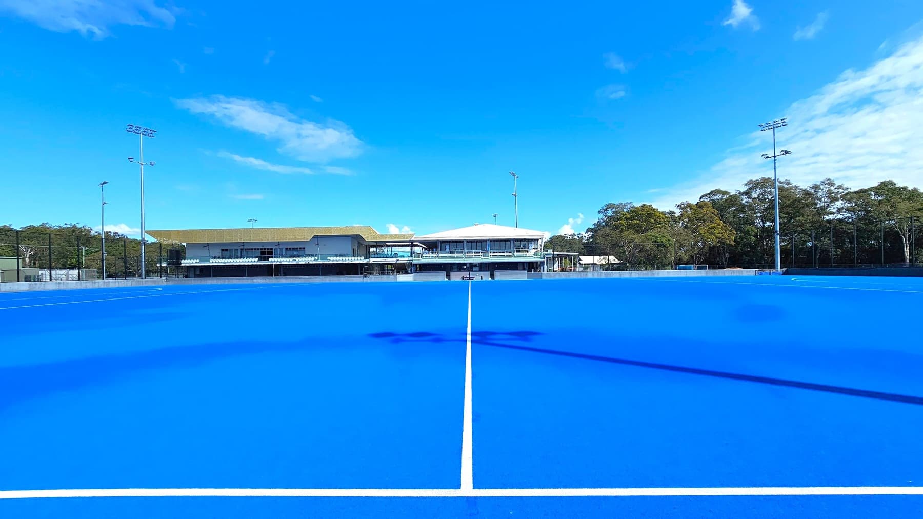A bright blue hockey field stretches across the foreground, with a white line running through it. In the background, the Gold Coast Hockey Centre building is visible, featuring a yellow roof and a row of seating. Tall floodlights are positioned on both sides of the field. Trees and a few small structures are visible to the right, while the left side shows more trees and a parked vehicle. The sky is partly cloudy.