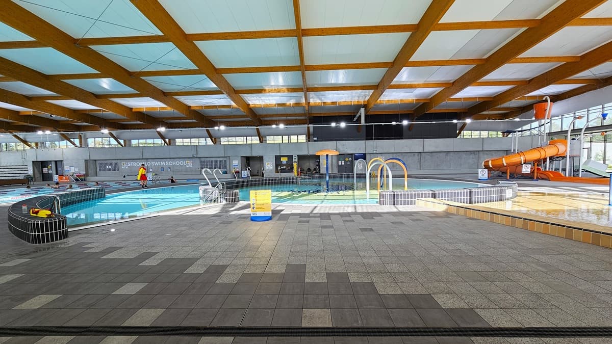 The children's splash park and leisure pool include a beach entry, water sprays, geysers, and waterfalls. The pool features a raised deck with small black tiles, bordered by grey checkered tiles. A yellow safety sign is visible. An orange slide with a tipping bucket is situated on the right, near the glass wall. The ceiling is adorned with wooden beams, and a pool staff member is stationed beside the pool.