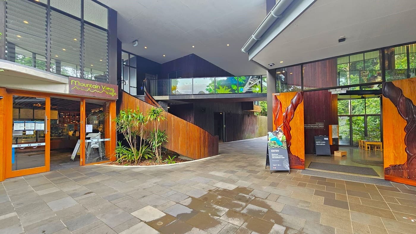 On the left, there is the entrance to the Mountain View Café with wooden doors and a sign above. Inside, a counter and menu are visible. In the centre, a staircase with wooden railings leads to an upper level with glass panels featuring nature-themed designs. On the right, large wooden doors with carved details open into a space with a sign about entry donations. The floor is paved with multicoloured tiles, and there are plants near the café entrance.
