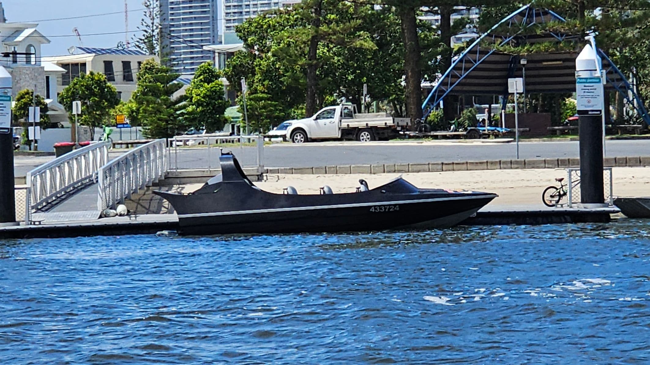A sleek black speedboat is moored to a concrete dock on the right. The boat has a low profile with grey seating. In the background, a sandy beach is lined with tall pine trees and several modern high-rise buildings. On the left, the water stretches out with numerous sailboats anchored in the distance. A black and yellow striped post is visible on the dock.