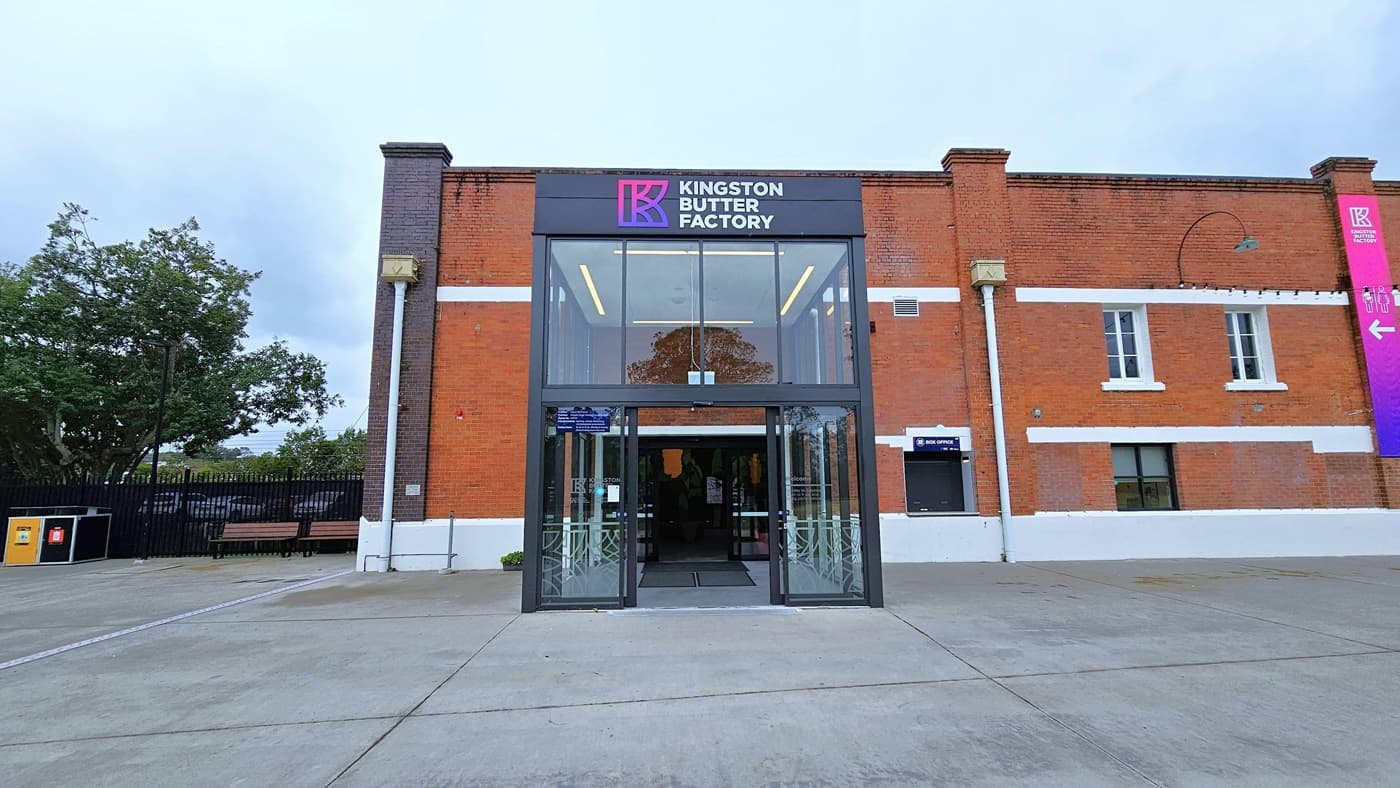 In the middle stands an industrial building made of red bricks with glass windows. Its front is adorned with a gradient blue and pink banner, vertically hung on the right side along with light fixtures. The entrance, a double modern glass and steel structure, features facility signage atop the glass door. On the left, a black steel gate and rubbish bins are present. Trees are also within the fenced area.