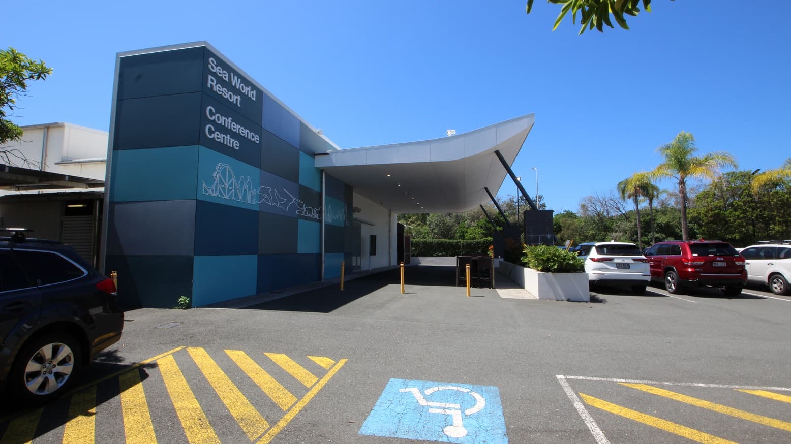 Drop off point for the Sea World Resort Conference Centre showing the 3 accessible car parks on the left hand side of the facility.  The conference centre is a single story building with large grey and blue tiled exterior.  A large portico offers under cover drop off areas.