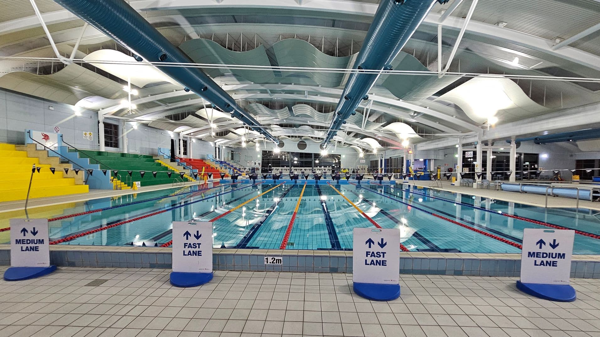 An indoor swimming pool with multiple lanes, each marked by floating lane dividers. Signs at the pool's edge indicate "Medium Lane" and "Fast Lane". On the left, there are tiered seating areas with yellow, green, and red seats. The ceiling features large, curved panels and exposed ductwork. The floor is tiled in light colours, and the pool depth is marked as 1.2 metres. Bright overhead lighting illuminates the space.