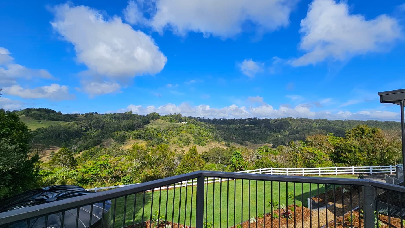 On the left, there is a black railing overlooking a green landscape with trees and hills. In the middle, a white fence runs along the edge of a well-manicured lawn. On the right, part of a building with a grey roof and a black railing is visible. The foreground features a grassy area with a small garden bed. The floor is a mix of grass and mulch, and there is no visible ceiling.