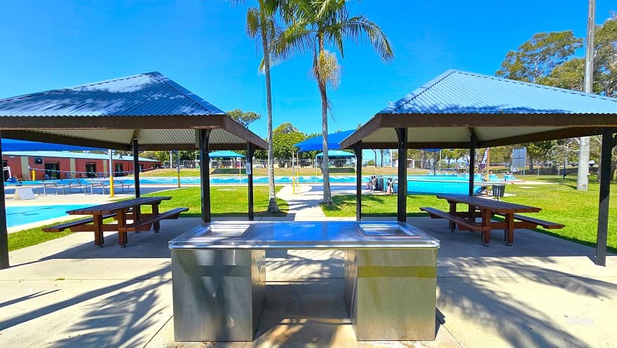 View in front of the stainless steel outdoor BBQ at Deception Bay Swimming Pool. Two park shelters with corrugated roofing with dark brown roof supports. Each shelter has a wooden picnic table. The outdoor BBQ is next to the splash park and coconut trees that partially shade the BBQ grilling station.