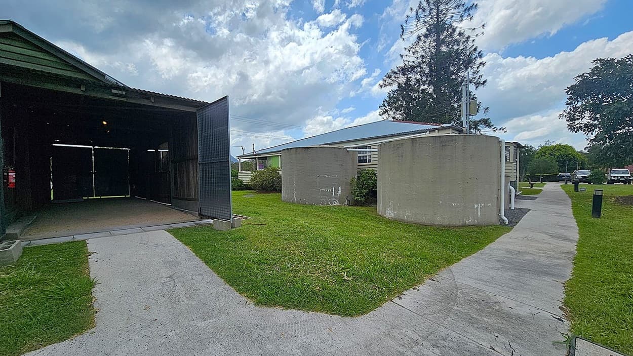 A concrete path splits into two directions. On the left, there's a dark, open shed with a corrugated roof and a fire extinguisher mounted inside. On the right, two large cylindrical water tanks stand beside a building with a blue roof. The grass is neatly trimmed, and a few trees are visible in the background. A couple of parked cars can be seen further down the path.