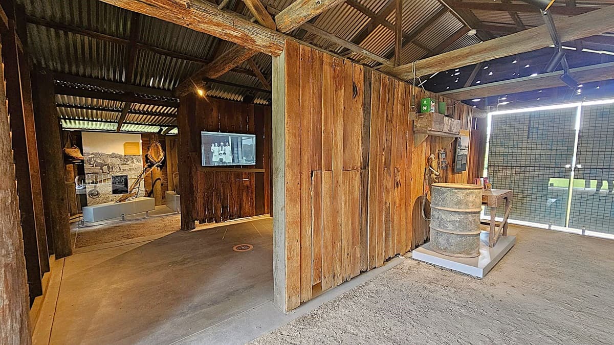 A rustic interior with wooden walls and a corrugated metal roof. On the left, there's a display area with historical photographs and artefacts, including a large rope and a plough. In the centre, a screen shows a black-and-white image. On the right, a metal barrel and a small workbench are placed on a platform. The floor is a mix of concrete and dirt, and the background features a mesh screen with a view of greenery outside.