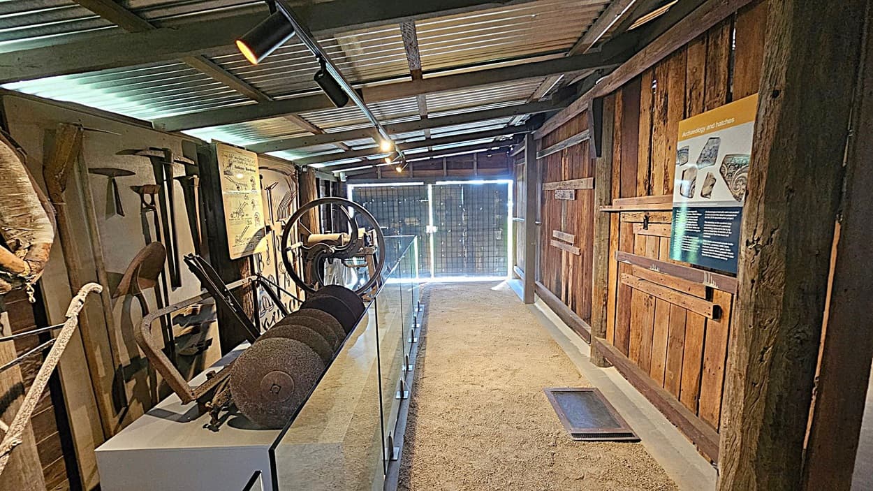 A narrow indoor space with a corrugated metal ceiling and a dirt floor. On the left, various old tools and machinery are displayed, including a large wheel and grinding stones. The right side features a wooden wall with a poster about archaeology and hatchets. At the end of the room, a glass barrier and a metal gate allow light to enter. Ceiling lights illuminate the area.