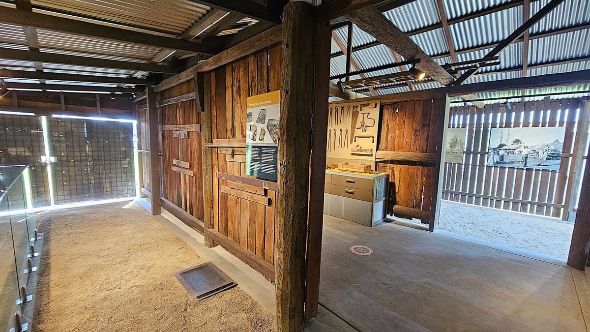 A rustic interior with wooden walls and a corrugated metal ceiling. On the left, a glass display case stands on a sandy floor. In the centre, a wooden partition features a poster about archaeology and artefacts. To the right, a room with tools displayed on the wall and a cabinet below. The floor is concrete, and a historical photograph is mounted on the wooden slatted wall, allowing light to filter through.