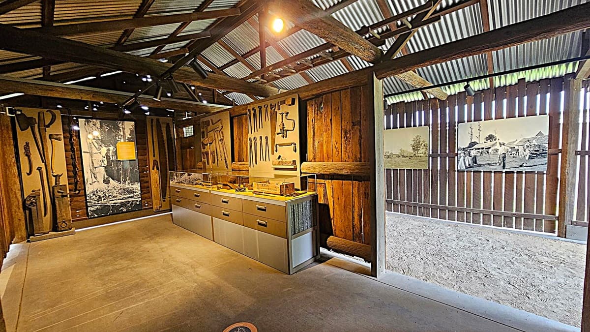 Inside a rustic wooden structure with a corrugated metal roof. On the left, a display of vintage tools is mounted on the wall, alongside a large black-and-white photograph. In the centre, a glass-topped display case with drawers underneath showcases more tools. The right side features two historical photographs on a slatted wooden wall, allowing light to filter through. The floor is concrete, and the ceiling is supported by wooden beams.