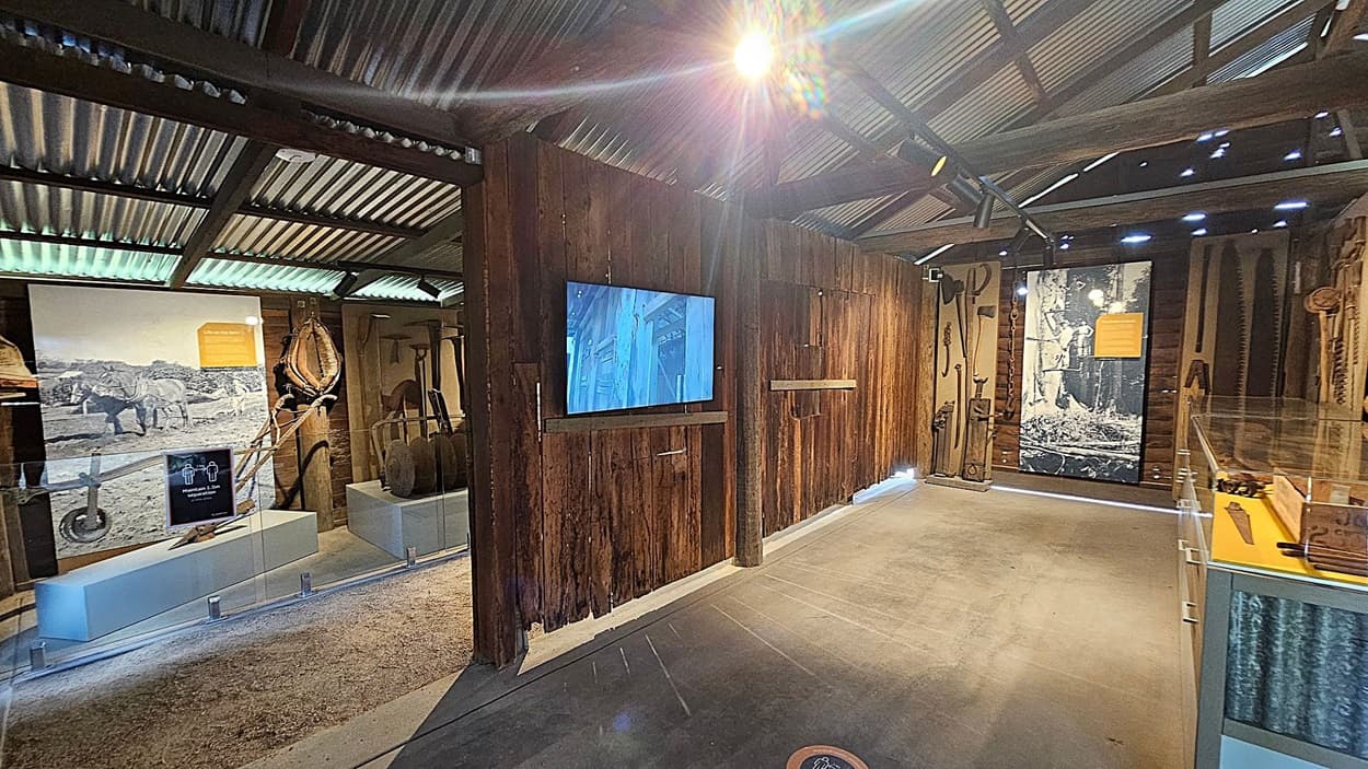 A museum exhibit with a rustic theme. On the left, there are displays featuring historical tools and equipment, including a large photograph of horses pulling a plough. A wooden wall in the centre has a mounted screen showing a video. On the right, more tools are displayed against a wooden backdrop, with a glass case containing additional artefacts. The floor is concrete, and the ceiling is made of corrugated metal, adding to the industrial feel.