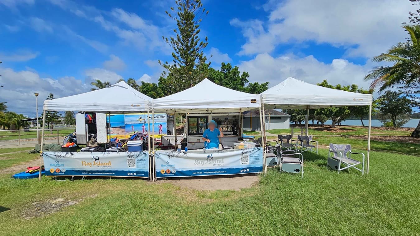 A grassy area with a trailer on the left displaying "Bay Island Aqua Accessibility" and kayaks nearby. In the centre, there are three white tents with banners and equipment. On the right, a blue beach wheelchair with large wheels is visible. Trees and a glimpse of water are in the background, with a mix of palm and other trees. The sky is partly cloudy.