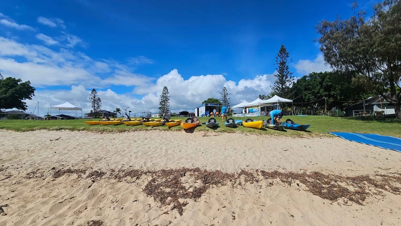 A sandy beach in the foreground leads to a grassy area where several kayaks are lined up. On the right, a person is adjusting a kayak. In the background, there are white tents and a few buildings, with tall trees scattered around. A blue mat is laid out on the grass to the right.