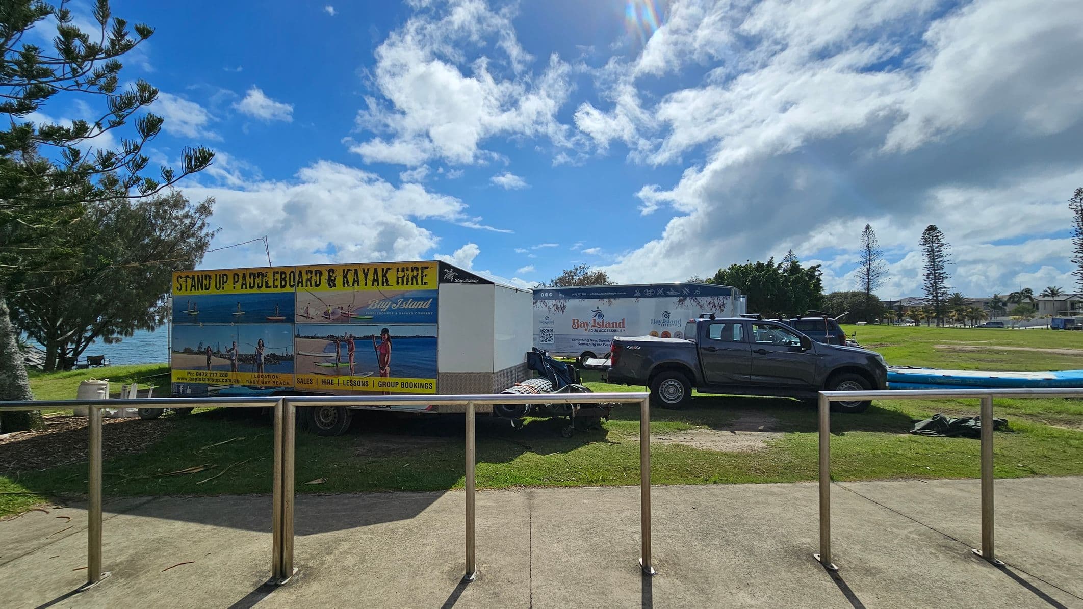 A grey pickup truck is parked on grass next to a trailer with a sign advertising "Stand Up Paddleboard & Kayak Hire." The trailer features images of people paddleboarding. In the background, there are trees and a glimpse of the sea to the left. A metal railing runs along the foreground on a concrete path. The sky is partly cloudy, and there are some tall trees in the distance on the right.