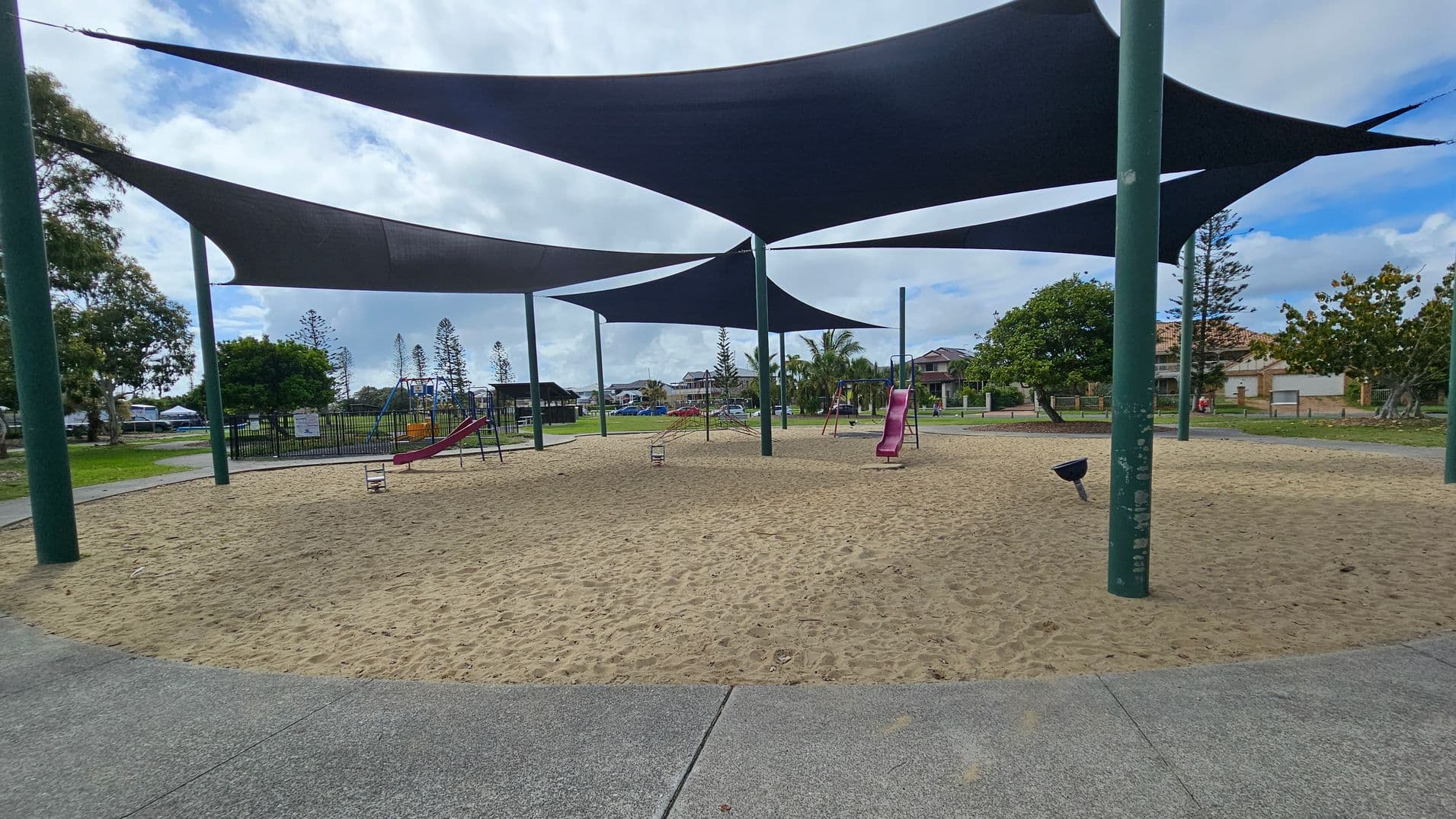 A park with a playground in the centre, featuring slides and swings under large shade sails. The ground is covered with sand. A concrete path curves through the park, with grass and trees on either side. On the right, there's a bench and more trees. In the background, houses are visible beyond the park.