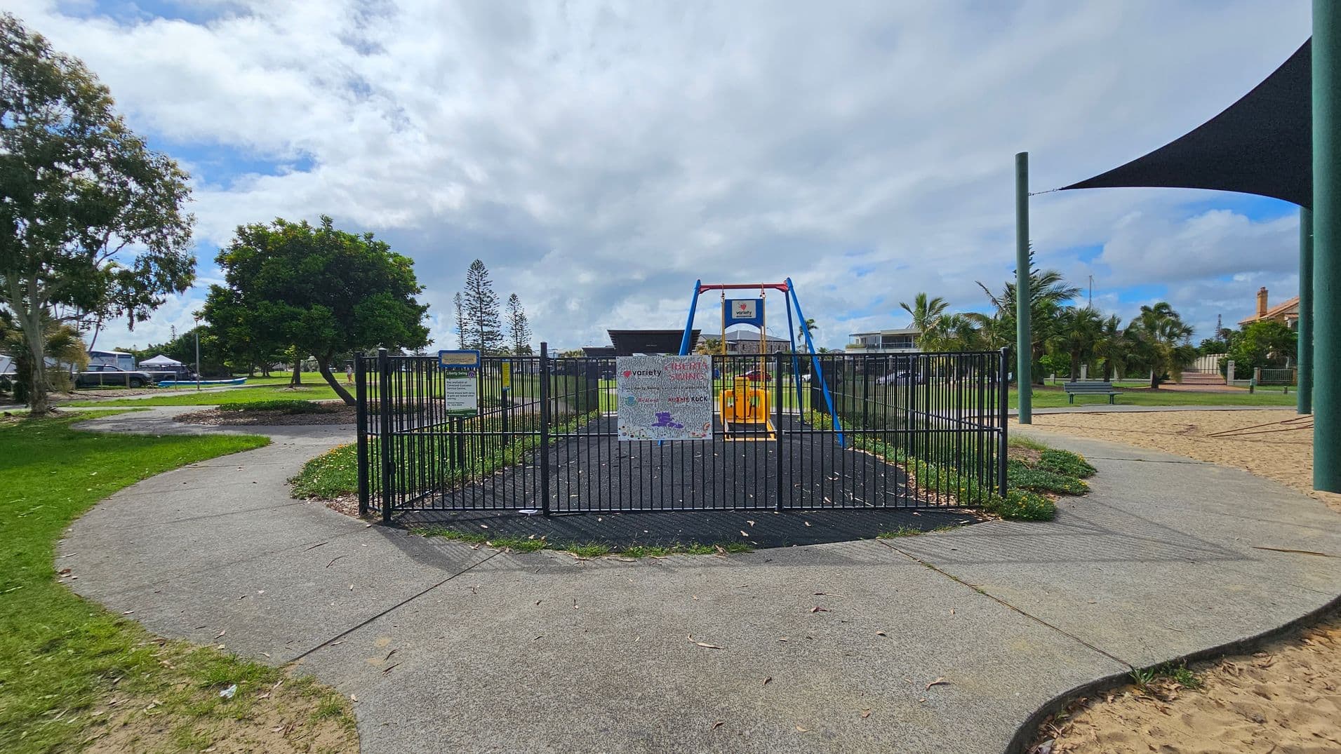 A fenced playground with a swing set in the centre, featuring a blue and red frame and a yellow seat. The ground inside the fence is covered with dark rubber matting. A sign with various logos is attached to the fence. Surrounding the playground is a paved path, with grass and trees on the left. On the right, there are palm trees, a bench, and a sandy area with a large triangular shade sail supported by green poles.