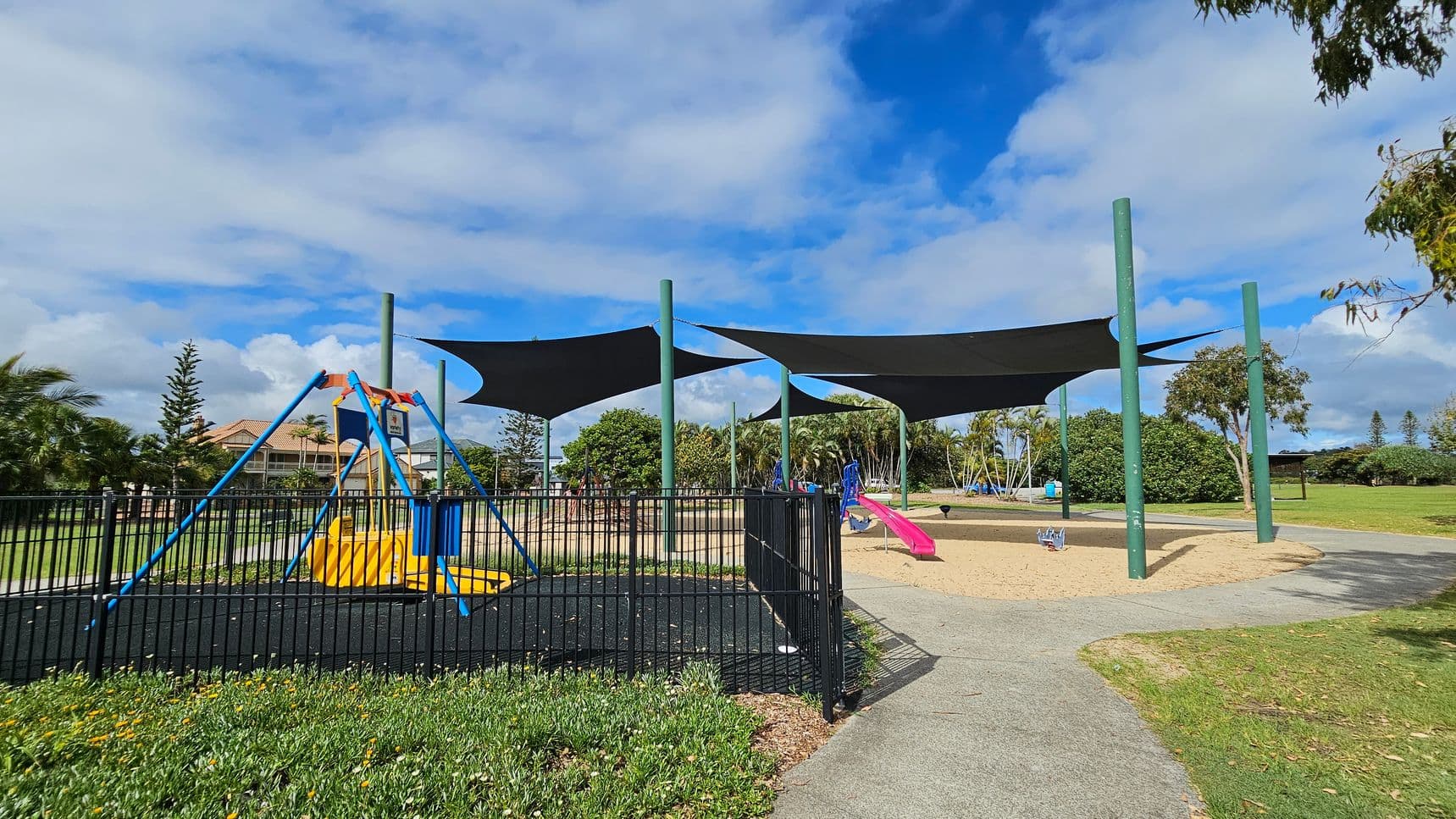 A playground with a fenced area on the left featuring a blue and yellow swing set. In the centre, there are large black shade sails supported by green poles. To the right, a pink slide sits on a sandy surface. The background includes trees, bushes, and a house with a tiled roof. The ground is a mix of grass and paved paths.