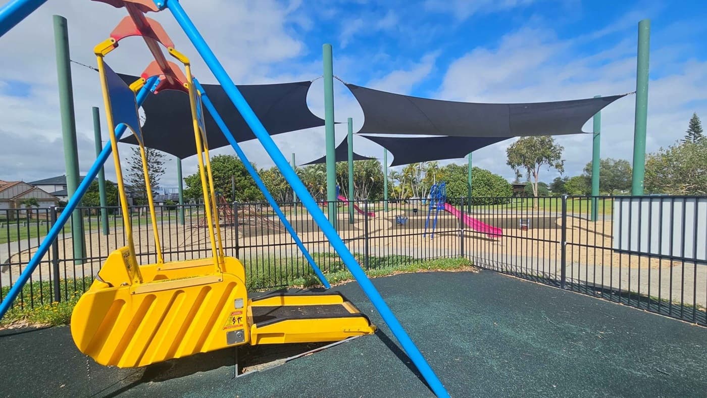 A playground with a yellow swing designed for accessibility on the left, supported by blue poles. The ground is covered with a dark rubber surface. In the background, there are several large, dark shade sails supported by green poles, providing cover over a sandy play area. A pink slide and other playground equipment are visible. The area is enclosed by a black metal fence, with trees and bushes in the distance.