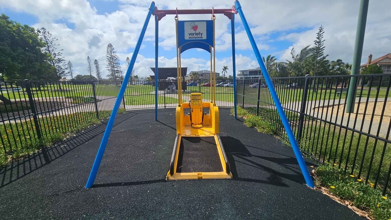 A playground swing designed for accessibility, featuring a yellow seat with a ramp for wheelchair access. The swing is supported by blue metal frames and is set on a black rubber surface. In the background, there is a grassy area with trees and a few buildings. A black metal fence surrounds the playground area.