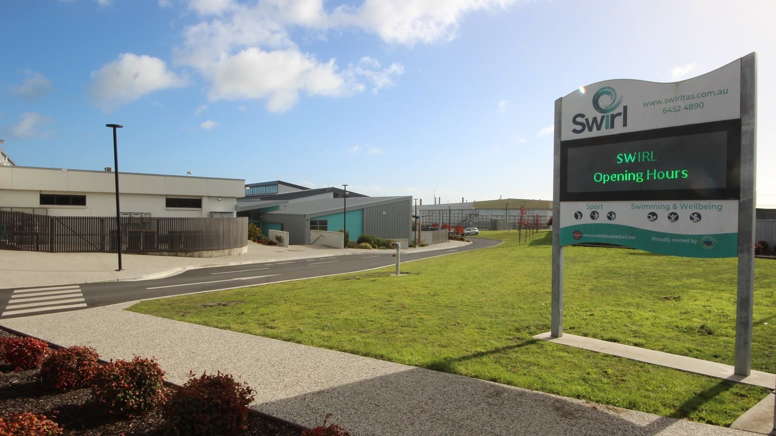 There are five accessible parking spaces near the entrance, each clearly marked with blue wheelchairs and yellow diagonal markings. Yellow bollards are also in place. The building on the right has alternating colors of green and grey. To the right is the entrance, featuring a huge "Barossa Aquatic Fitness" sign on the wall. A barricaded section with a water meter and controls is situated on the right.