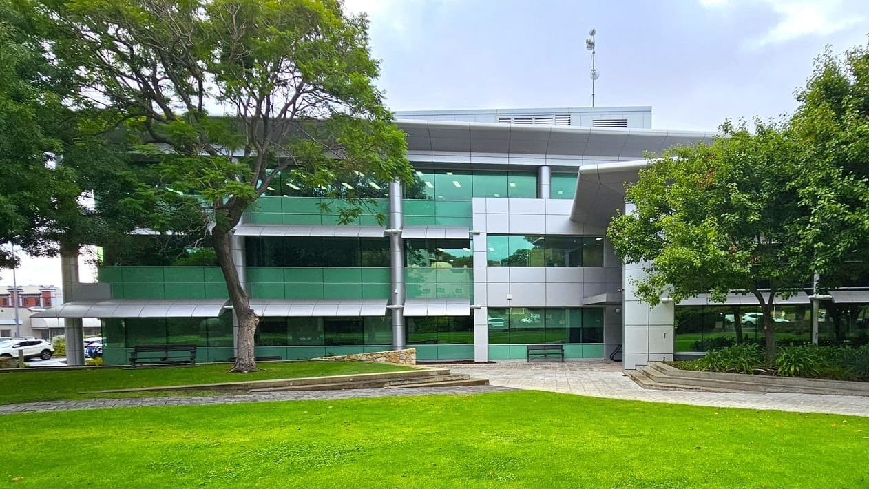 A modern building with large green-tinted windows and a grey facade is in the background. In the foreground, there is a well-maintained lawn bordered by a paved walkway. Trees with dense foliage are on both sides, providing shade. Benches are placed near the building, and there are landscaped areas with plants. The scene is calm and organised, with a mix of natural and architectural elements.