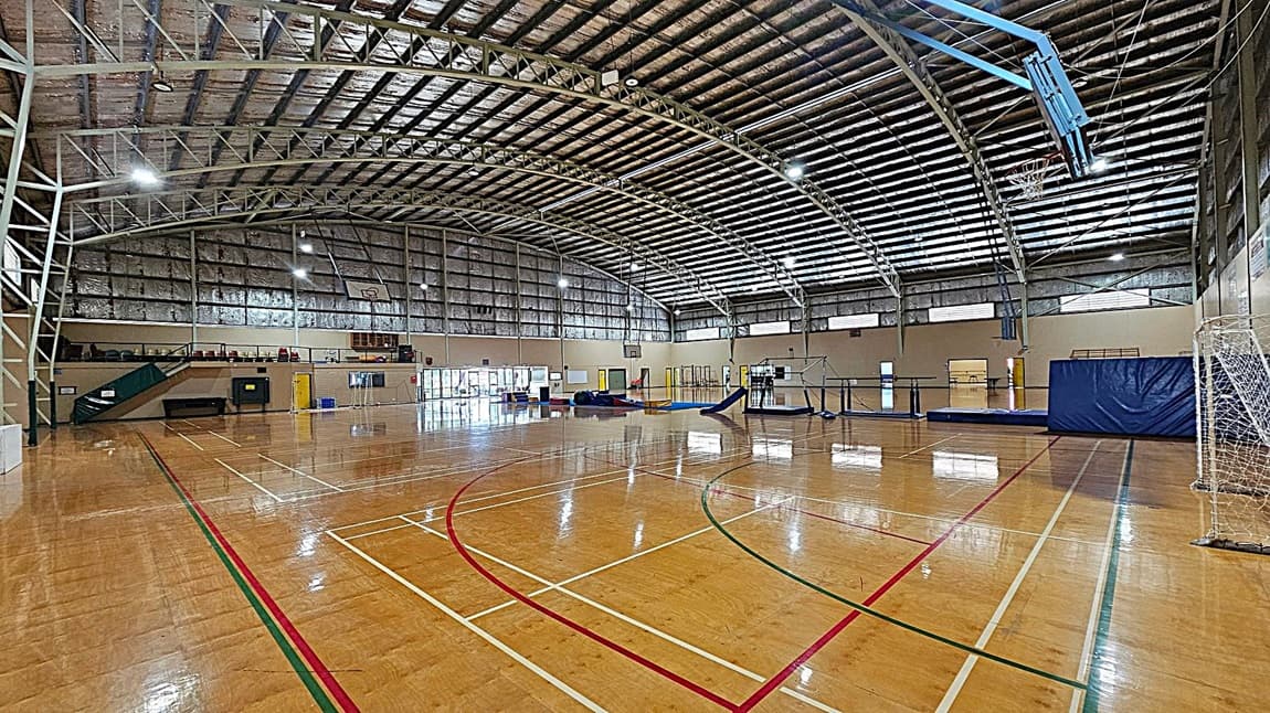 A large indoor sports hall with a high, arched ceiling made of metal beams and insulation. The polished wooden floor has various sports markings in red, green, and white. On the right, there's a basketball hoop and a netted goal. In the centre, gymnastic equipment is set up, including mats and beams. The left side features a staircase leading to a mezzanine with seating. The walls have several doors and windows, allowing natural light to enter.