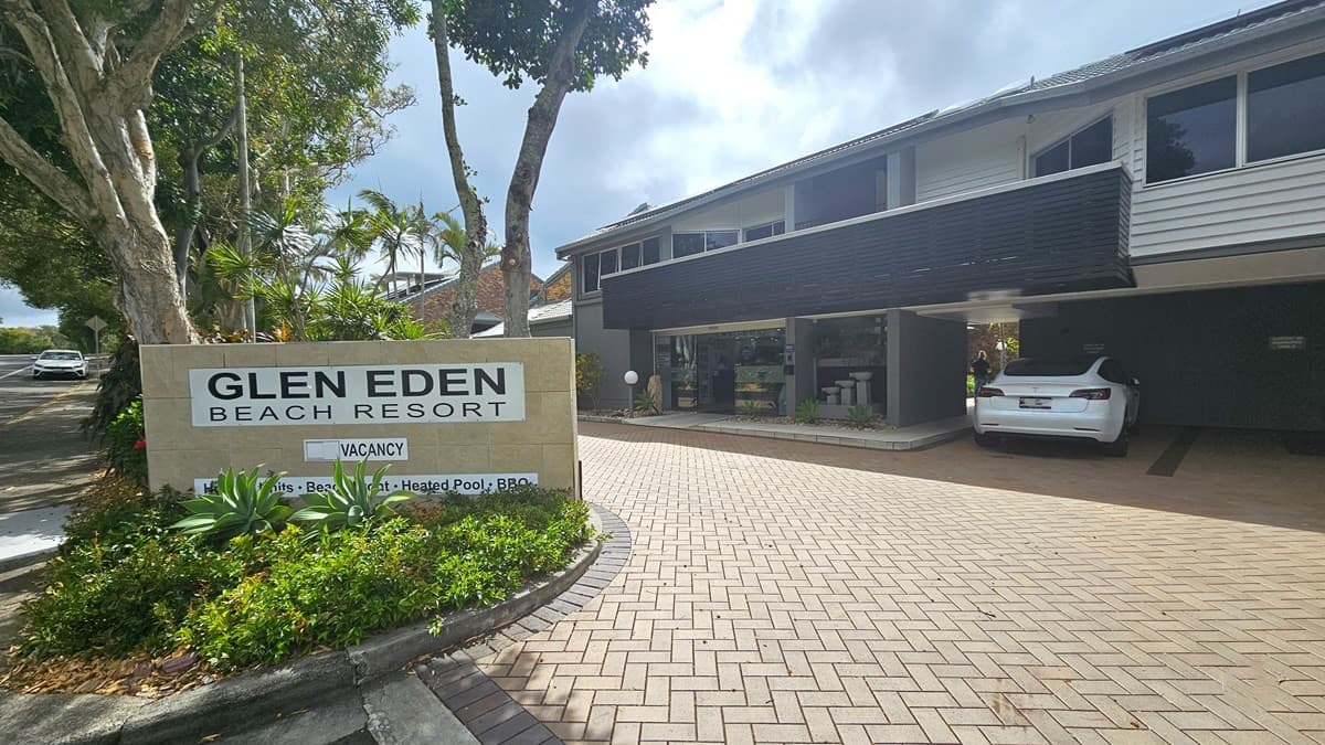 To the left is a road and to the right is a two-storey building with brown and white hues, accented by timber elements and grey corrugated iron roofing. Brick pavers pave the surrounding area. Signage flanks the entrance, amidst landscaped plants and trees. A white vehicle is parked at the undercover parking at the right of the entrance.