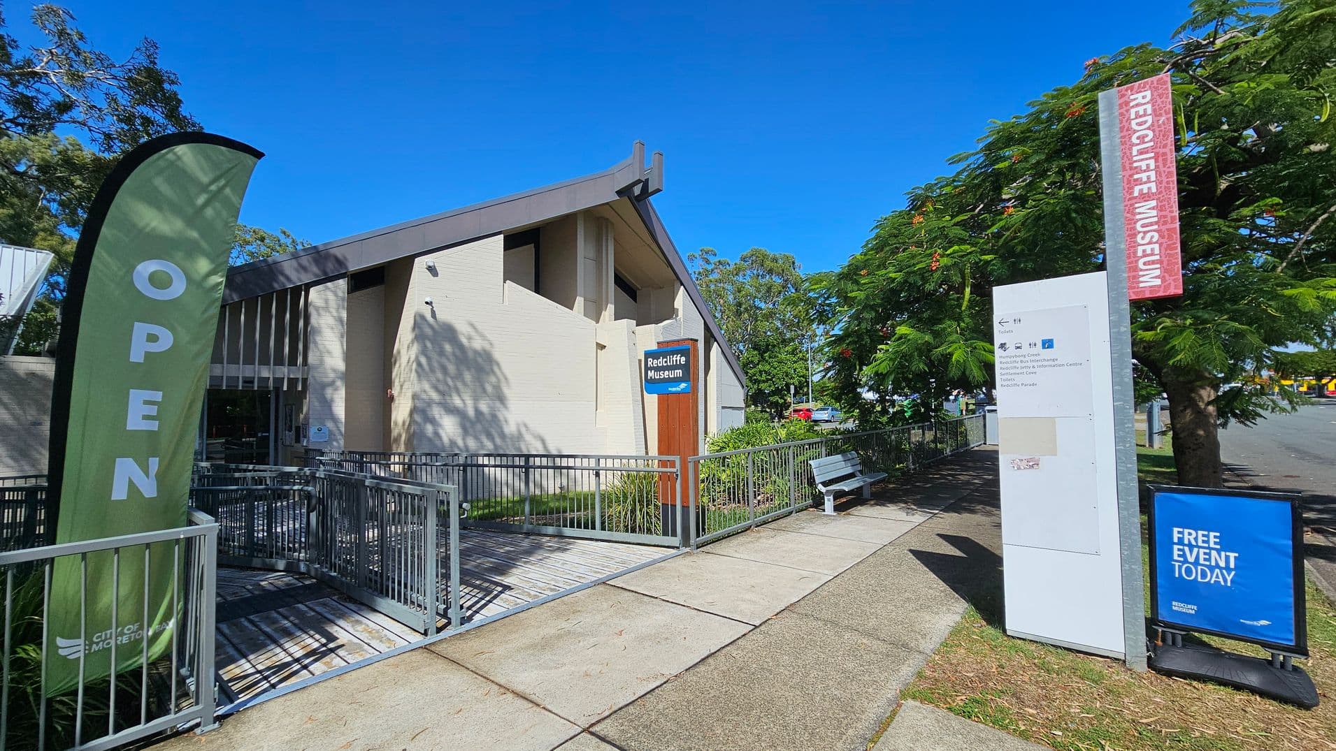 A building with a slanted roof and a sign reading "Redcliffe Museum" is on the left. A green flag with "OPEN" is near the entrance, which is accessible via a ramp with metal railings. On the right, a tall signpost and a blue sign advertising a "FREE EVENT TODAY" are visible. A bench is situated under a tree with red flowers. The pavement is a mix of concrete and grass, and the sky is clear and blue.