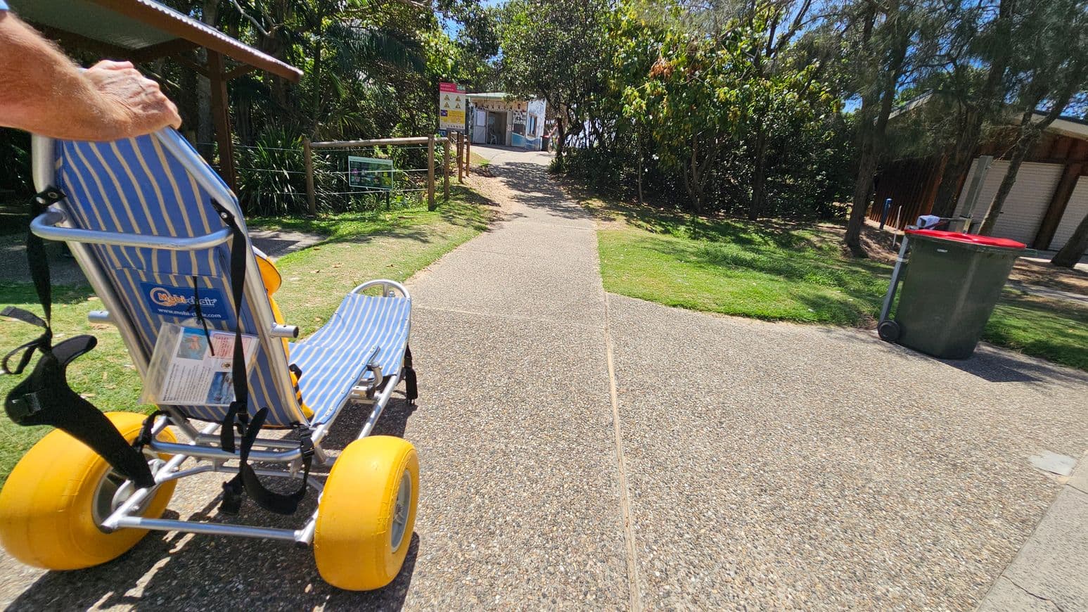 A blue and white striped beach wheelchair with large yellow wheels is on a textured pathway. To the left, there's a grassy area with kayaks in the background. A large tree stands in the centre, providing some shade. On the right, a path leads towards a wooden structure and more greenery.