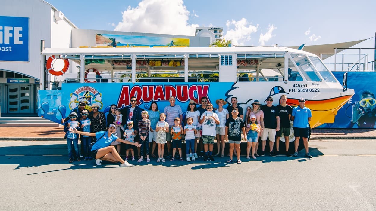 Group of happy adults and children posing for a photo in front of the Aquaduck parked near the red brick pathway and a blue fence that shows a professional diver with an okay sign and yellow diving goggles. Detailed below the driver’s right window are “Reggie 445751” and “PH: 5539-0222”. Further to the left is a white building with a blue sign that says “SEA LIFE Sunshine Coast” in white letters and a green perforated gate with a blue-green sign for ENTRY ONLY in white letters over above.