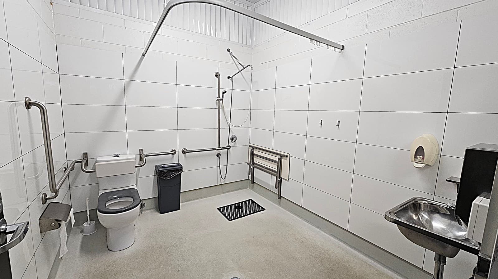 A spacious, accessible bathroom with white tiled walls and a light green floor. On the left, there is a toilet with grab bars and a toilet paper holder. A black bin is next to the toilet. In the centre, a shower area with a handheld showerhead and additional grab bars. On the right, a foldable shower seat is mounted on the wall. A stainless steel sink with a soap dispenser is also on the right. The ceiling is white with a curtain rail above the shower area.