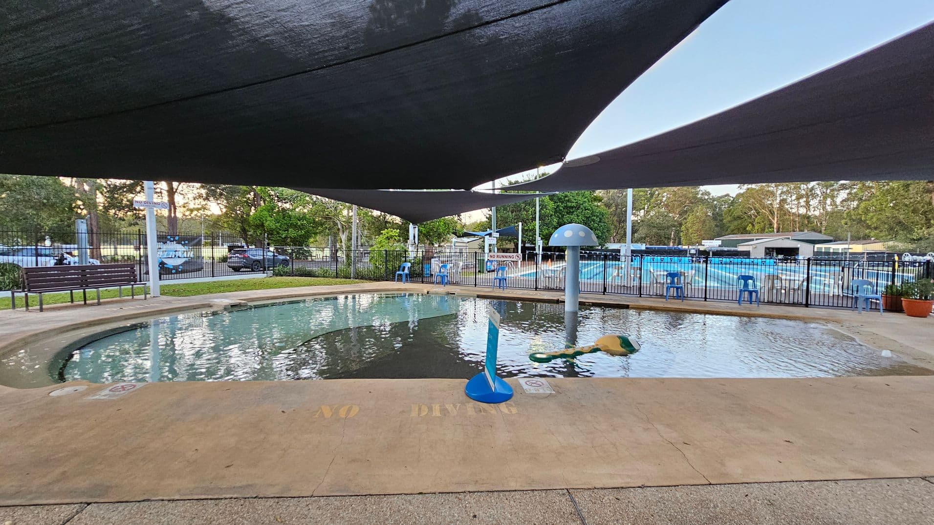 A small, shallow swimming pool is in the foreground, with a "NO DIVING" warning on the edge. A mushroom-shaped water feature is in the pool. Blue plastic chairs line the fence in the background, with a larger pool visible beyond. Shade sails cover the area above, and trees and parked cars are visible outside the fenced area. A bench is on the left side near the fence.