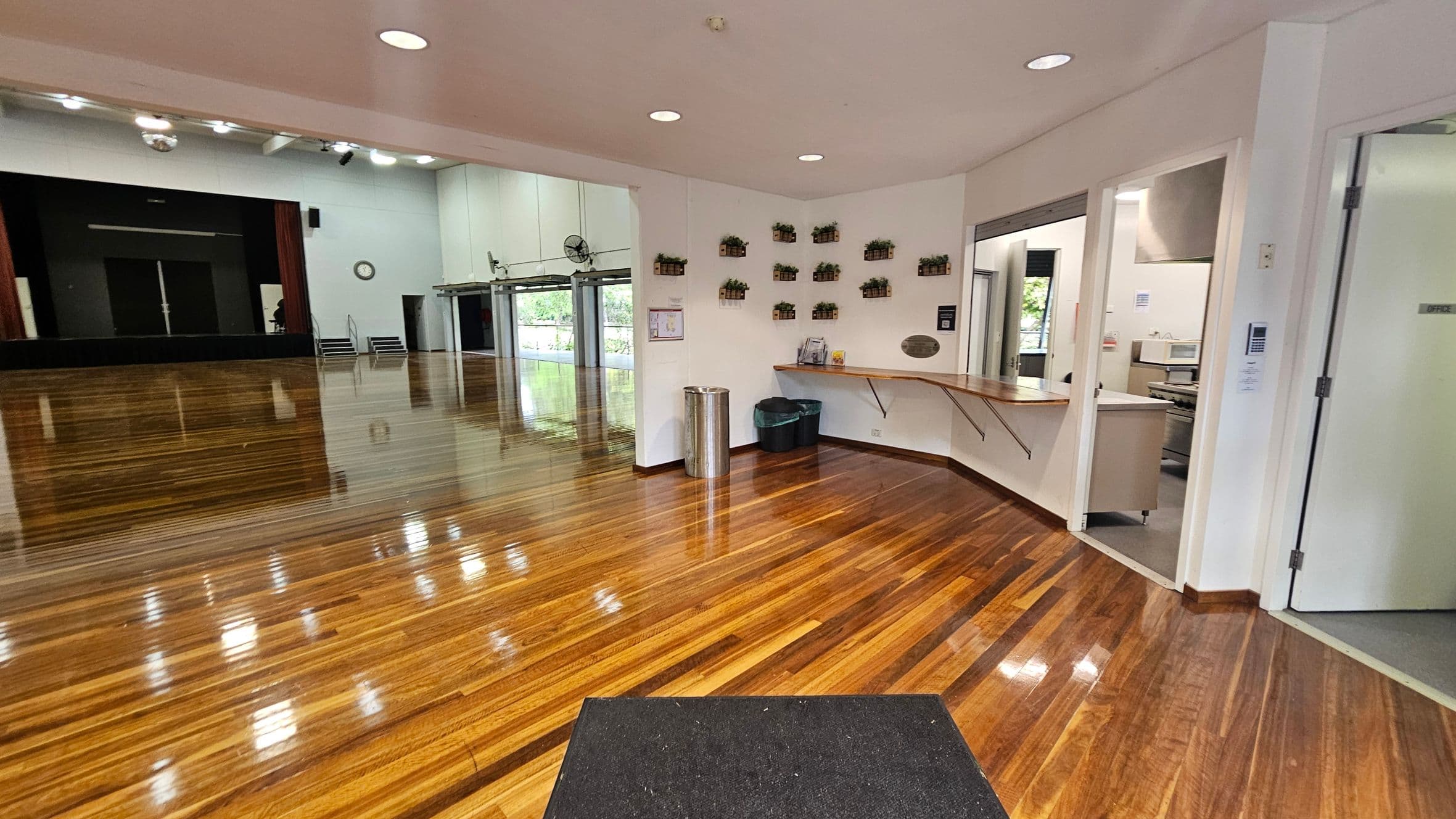 A spacious room with polished wooden floors. On the left, there's a large stage with a black backdrop and a clock on the wall. In the middle, a wall with small potted plants arranged in a pattern. On the right, an open doorway leads to an office area with a counter and a microwave. A black mat is on the floor near the entrance. The ceiling is white with recessed lighting.