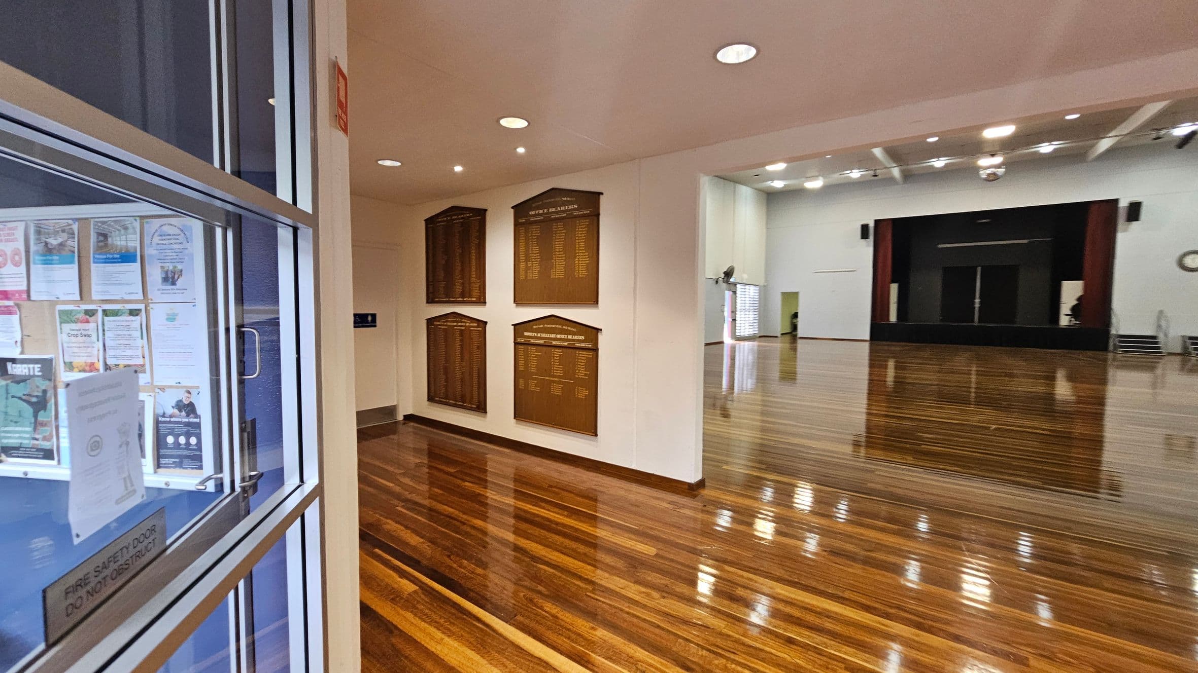 A spacious hall with polished wooden floors and a stage at the far end. On the left, a glass door with various posters and notices. The wall near the entrance displays several wooden plaques. The ceiling is fitted with recessed lights, and a disco ball hangs in the centre.