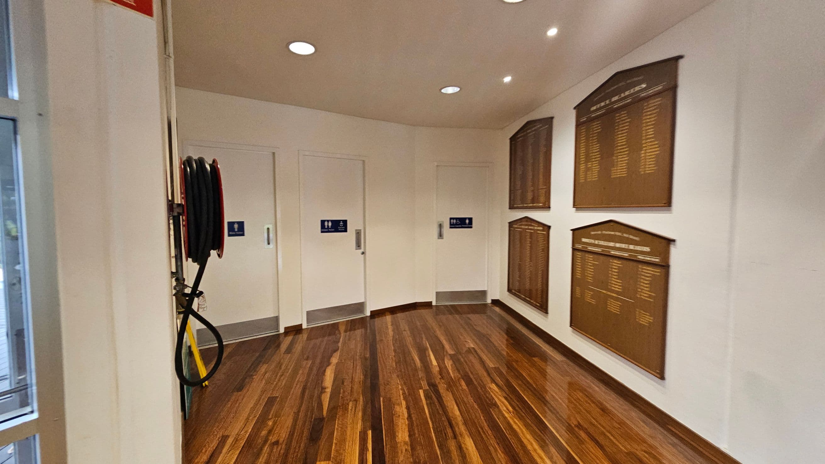 A hallway with polished wooden flooring and white walls. On the left, there's a fire hose reel mounted on the wall. In the centre, three white doors with blue signs indicating restrooms. On the right, several large wooden plaques with engraved text are mounted on the wall. The ceiling is white with recessed lighting.