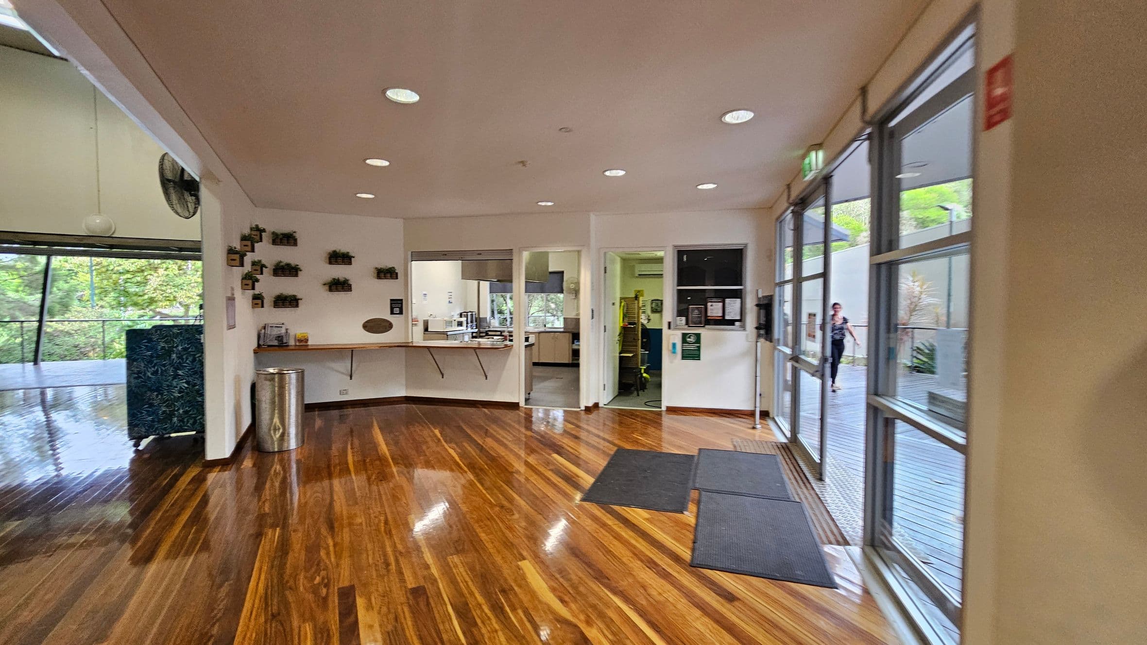 A room with polished wooden floors and a white ceiling with recessed lights. On the left, a wall with small potted plants and a counter with a bin beneath. In the centre, two open doorways lead to a kitchen area with visible appliances. On the right, large windows and glass doors open to an outdoor deck, where a person is walking. A couple of dark mats are placed on the floor near the entrance.