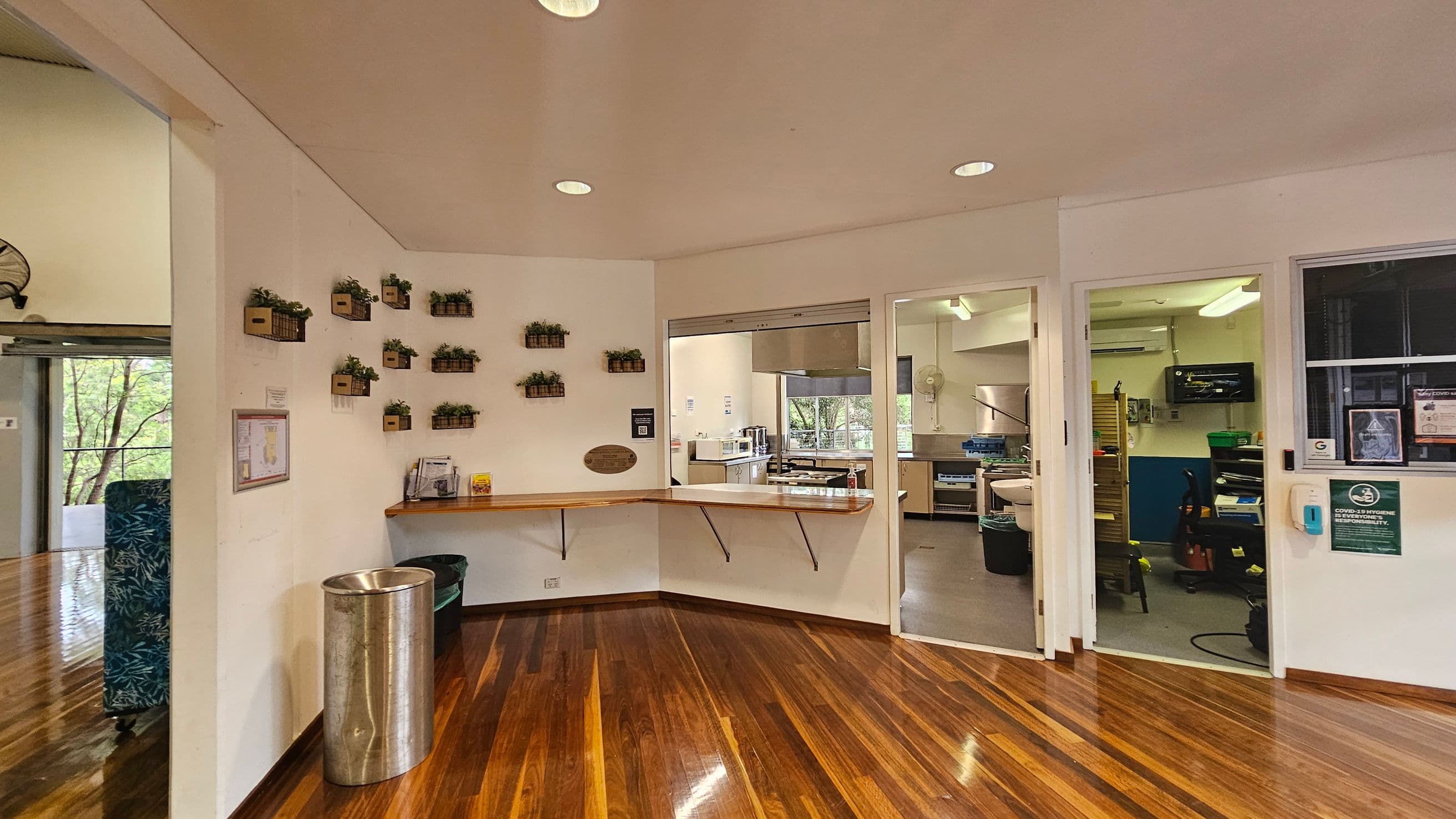 A room with polished wooden floors and a white ceiling. On the left, a wall with small potted plants arranged in a grid pattern. Below them, a wooden counter with brochures and a metal bin. In the centre, a window opens to a kitchen area with stainless steel appliances and a view of trees outside. On the right, a doorway leads to another room with office equipment and a wall-mounted screen. A hand sanitiser dispenser is mounted on the wall near the door.