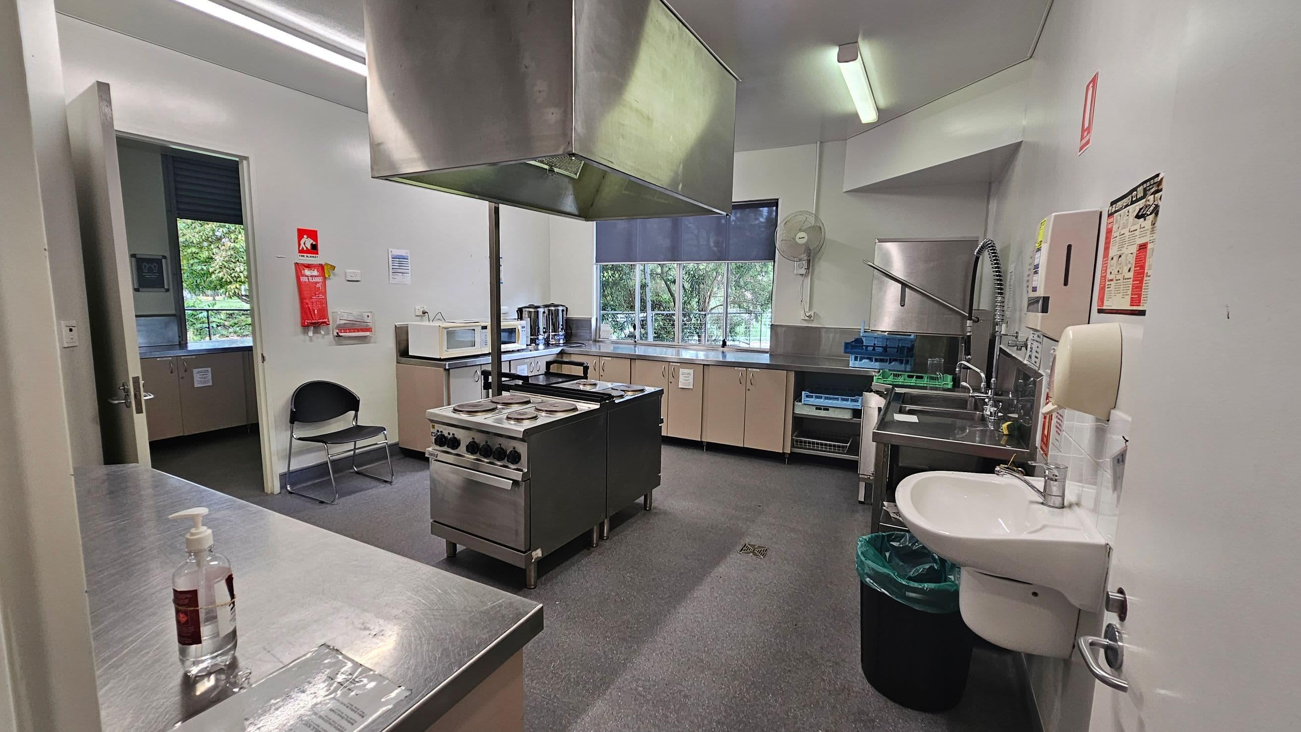 A commercial kitchen with stainless steel surfaces. On the left, there's a door leading to another room, a chair, and a microwave on a counter. In the centre, a large stove with an overhead extractor hood. On the right, a sink with a high faucet, a green bin, and wall-mounted dispensers. The back wall has a window with a view of trees, and cabinets below. The floor is grey, and the ceiling has fluorescent lights.