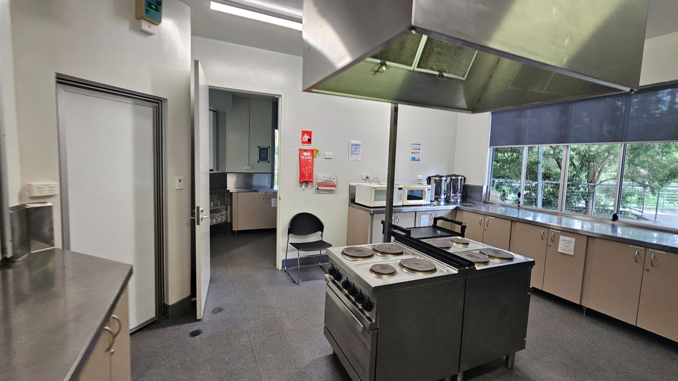 A commercial kitchen with a central stove featuring four burners and an overhead extractor hood. On the right, there are large windows with blue blinds, and a row of cabinets with a countertop holding microwaves and kettles. On the left, a door is open, leading to another room, and a fire extinguisher is mounted on the wall. The floor is grey, and the ceiling has fluorescent lighting.