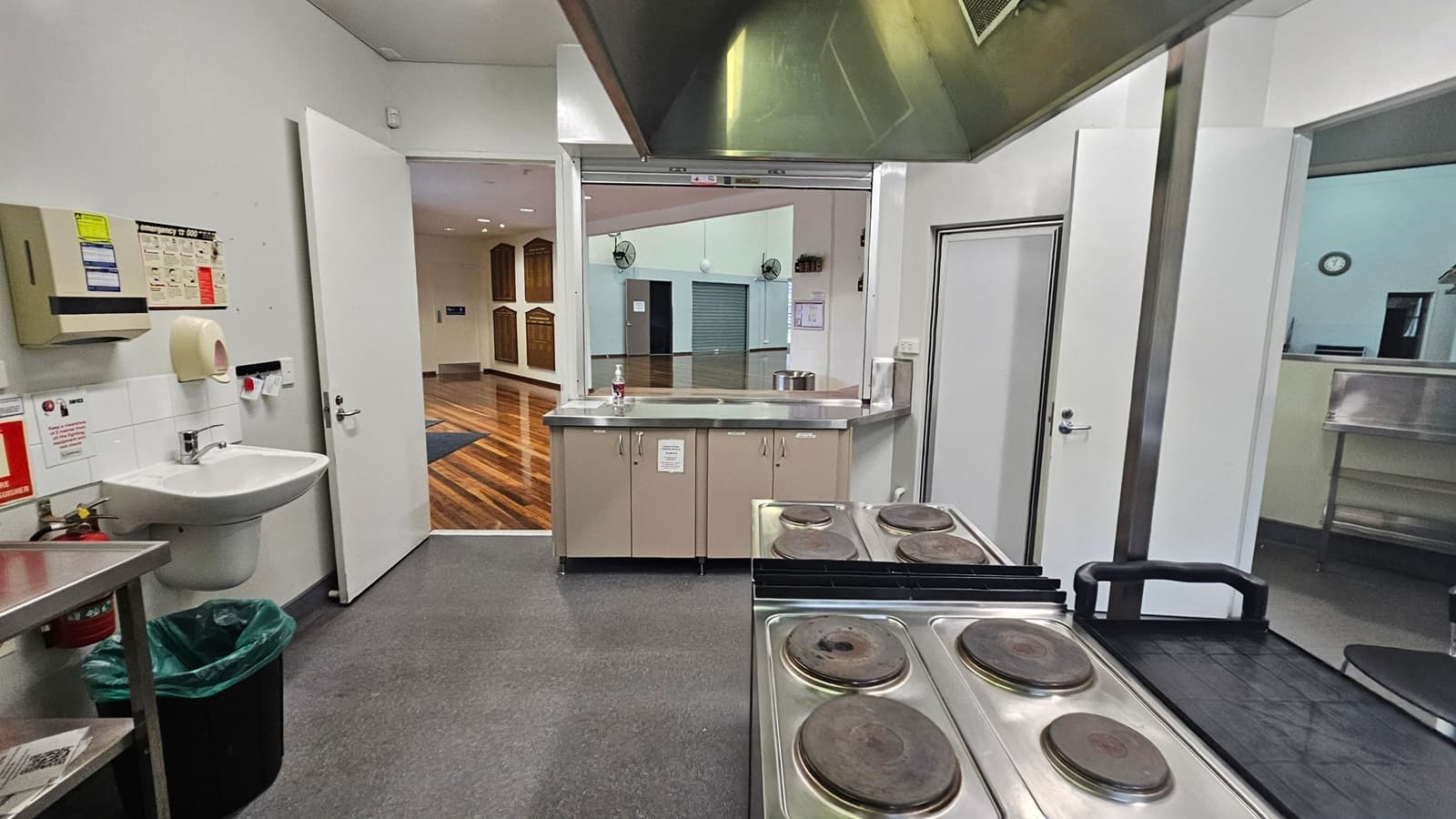 A commercial kitchen with a grey floor and white walls. On the left, there's a sink with a soap dispenser and a paper towel holder above it. A green bin is below the sink. In the centre, a counter with cabinets and a large pot is visible through an open serving hatch. The right side has a closed door and a stainless steel shelf. In the background, a room with wooden flooring and wall-mounted plaques is visible through an open doorway.