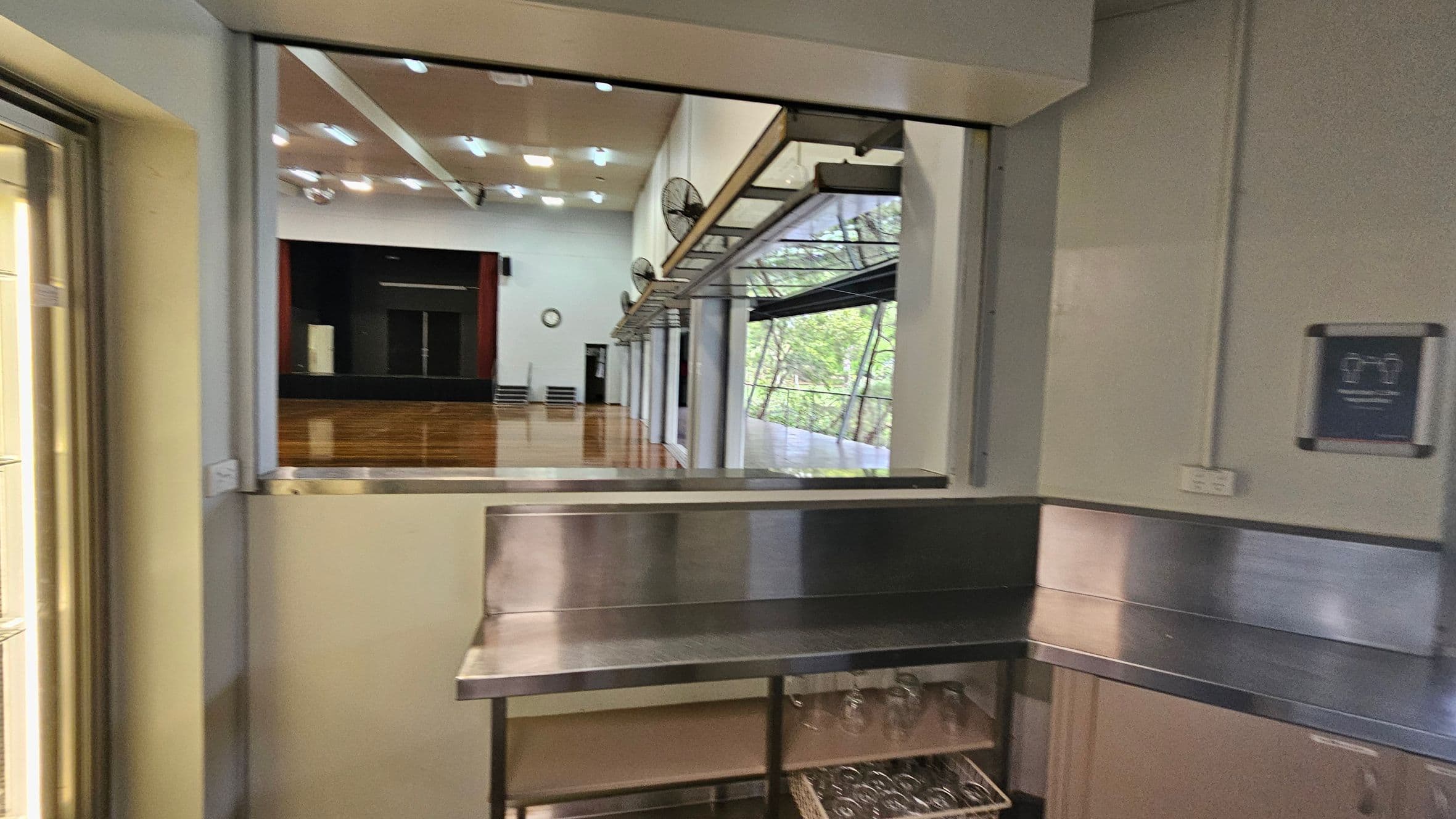 A view from a kitchen or serving area into a large hall. The foreground has a stainless steel counter with shelves below, holding glasses. The background shows a spacious hall with a wooden floor, a stage with red curtains, and a clock on the wall. Large windows on the right let in natural light, and ceiling lights are visible. A sign is mounted on the right wall.