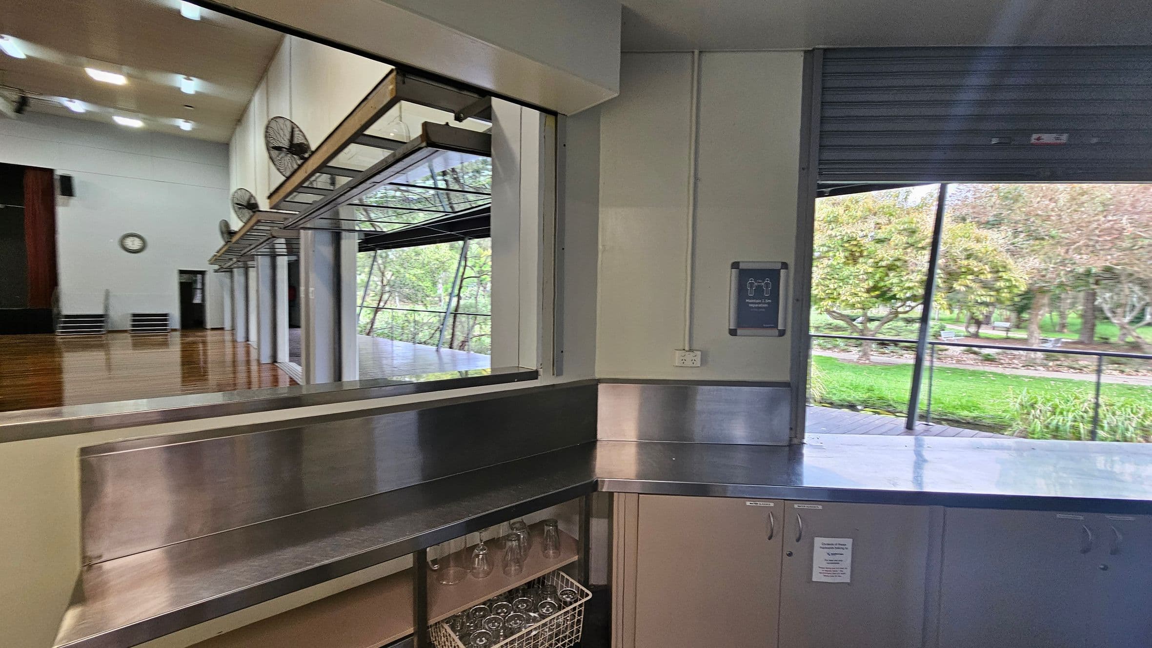 On the left, a large indoor space with wooden flooring and a wall clock. Ceiling fans are mounted above a serving counter with open windows. On the right, a stainless steel counter with a basket of glasses underneath. A sign is mounted on the wall above an electrical outlet. Through the open window, a view of a green outdoor area with trees and a pathway.