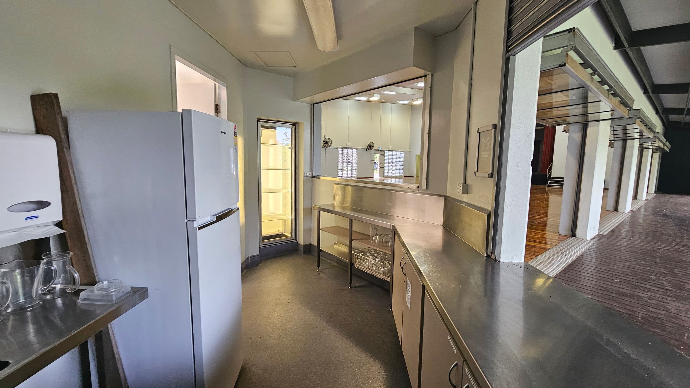 A small kitchen area with a white refrigerator on the left, next to a wall-mounted paper towel dispenser and some glass jugs on a metal counter. In the centre, there is a stainless steel counter with shelves underneath, and a window with a view into another room. On the right, a large open space with wooden flooring is visible through a wide serving hatch. The ceiling has a fluorescent light fixture, and the floor is covered with a dark, speckled material.