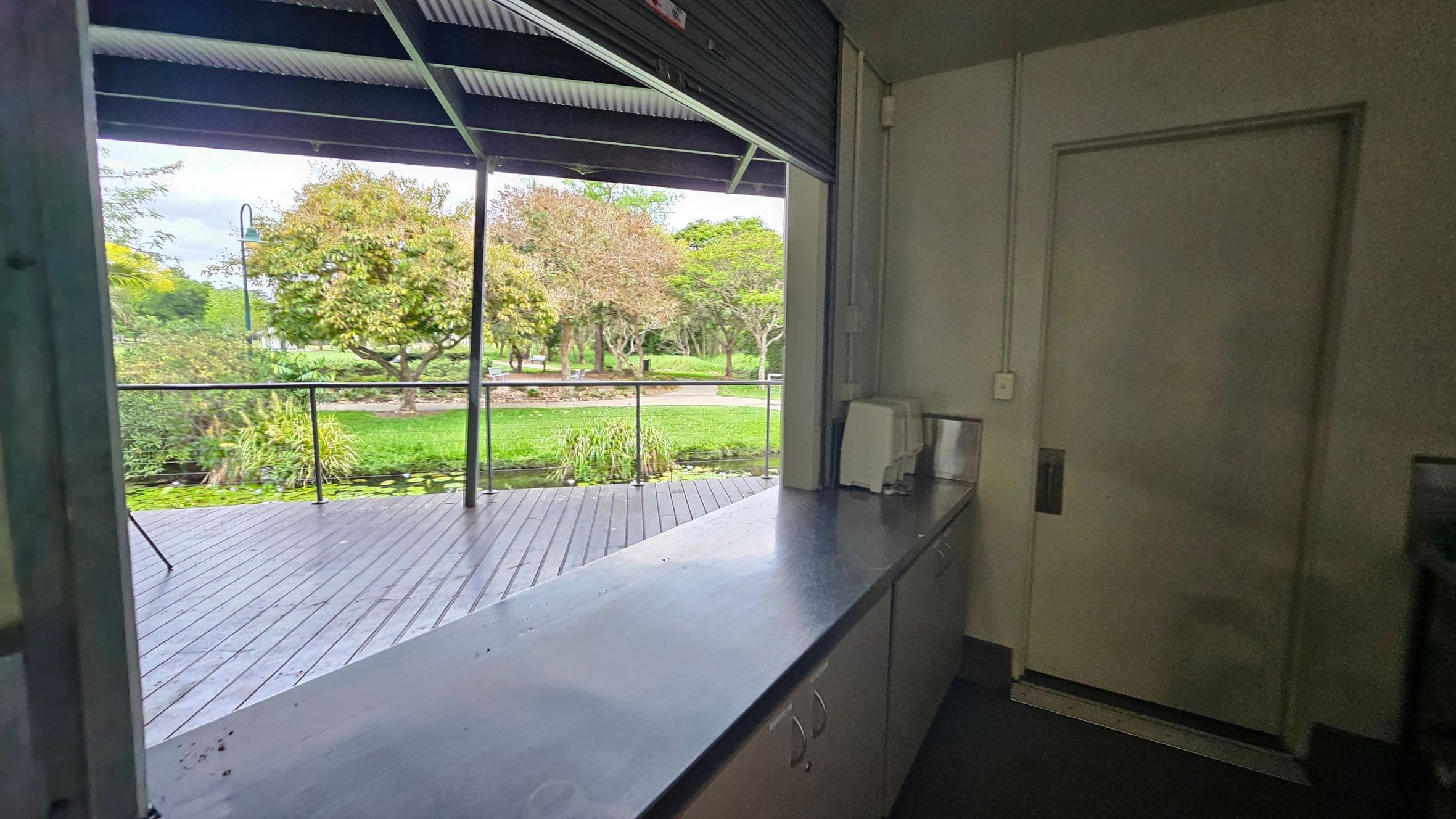 A view from inside a building with a metal counter on the right, leading to a partially open roller door. Outside, there is a wooden deck with a railing, overlooking a lush garden with green grass, trees, and a small pond with lily pads. A lamppost is visible among the trees. The ceiling inside is corrugated metal, and the interior walls are painted a light green.