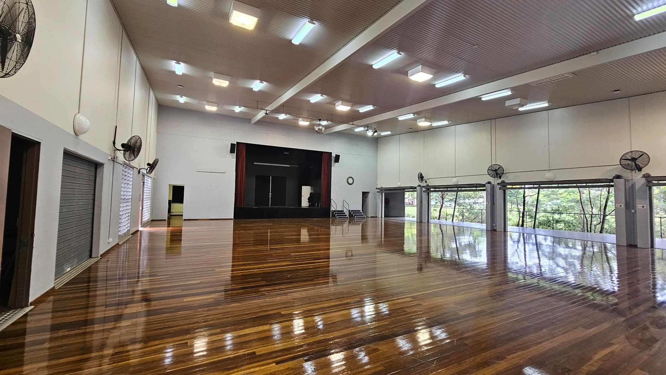 A spacious hall with a polished wooden floor and a high ceiling with fluorescent lights. On the left, there are large fans mounted on the walls and several closed roller doors. The back wall features a stage with dark curtains and steps leading up to it. On the right, large windows with views of greenery outside are visible, along with more wall-mounted fans. A clock is mounted on the wall near the stage.
