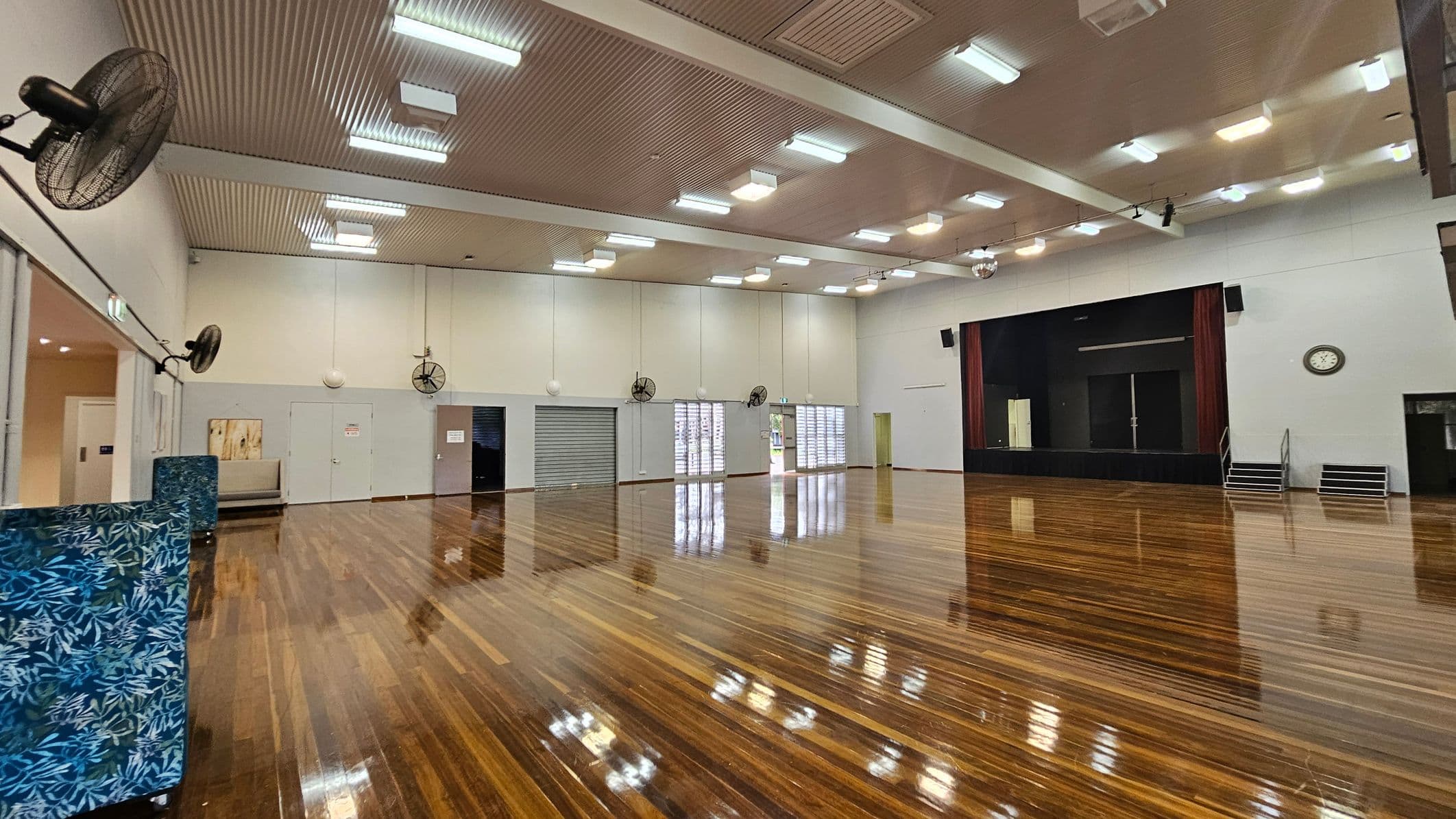 A large hall with polished wooden floors and a high, ribbed ceiling with fluorescent lights. On the left, there are wall-mounted fans and a small seating area with blue patterned dividers. The back wall features a stage with red curtains and a clock above it. Several doors and windows with shutters are visible along the walls. The right side has a set of steps leading to the stage. The space is open and well-lit.