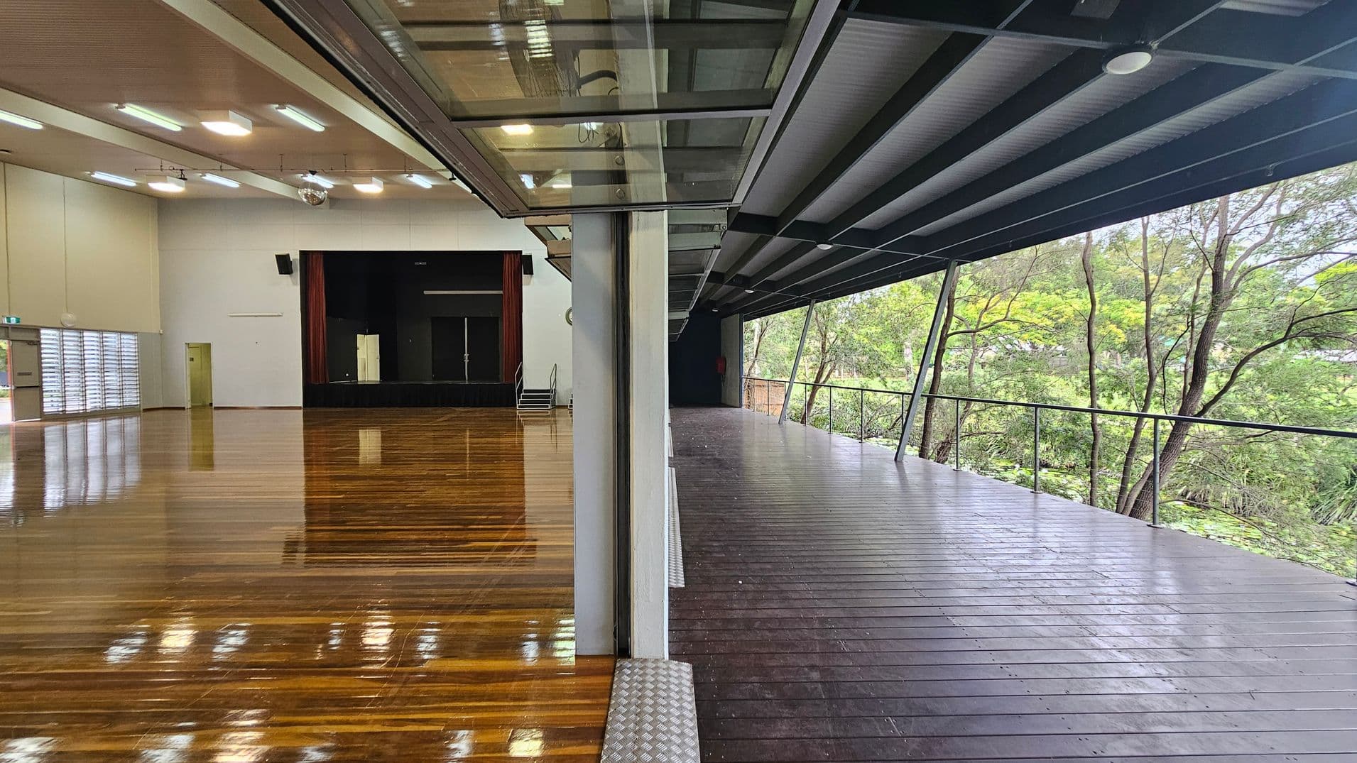 On the left, a large indoor hall with polished wooden floors and a stage at the far end. The ceiling has multiple fluorescent lights. On the right, an outdoor deck with wooden flooring and a metal railing, overlooking a lush green forest. The deck is partially covered by a roof with visible beams. A glass partition separates the indoor and outdoor spaces.