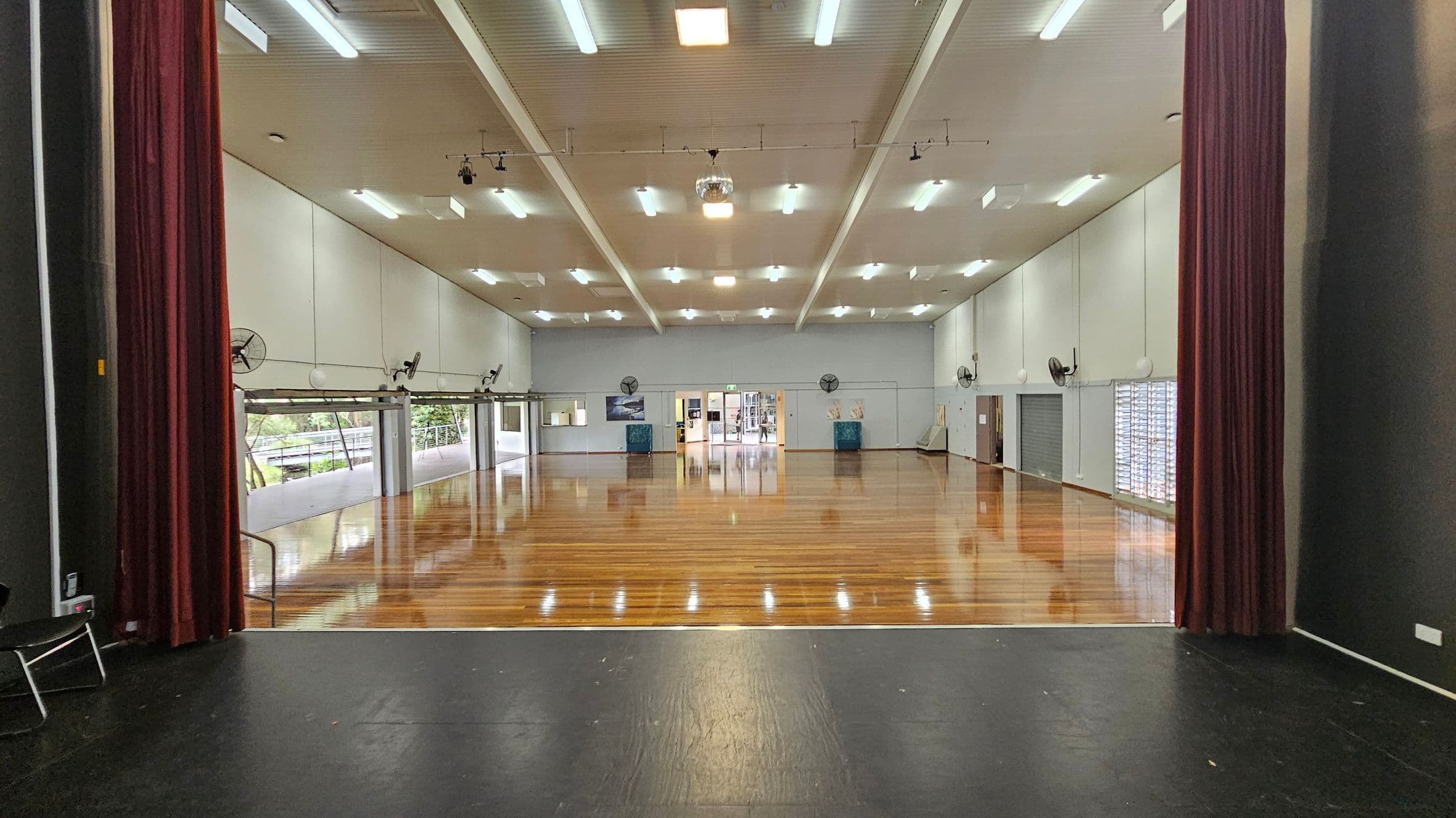 A large, empty hall with polished wooden floors and a high ceiling with fluorescent lights. The view is from a stage with dark flooring and red curtains on either side. The walls are light-coloured with several wall-mounted fans. At the far end, there are double doors leading to another room. Windows on the left side let in natural light, and there are more windows on the right with blinds partially drawn.