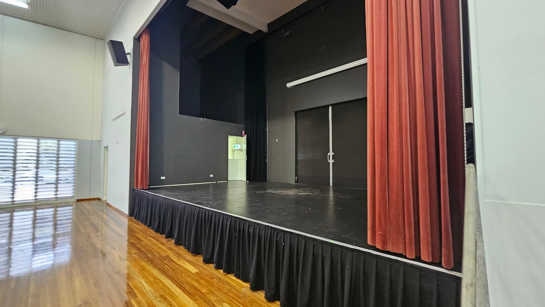 On the left, there are tall windows with horizontal blinds allowing light to filter through. In the middle, a black stage with red curtains on the right side and a white door slightly ajar, leading to a lit room. The floor is polished wood, reflecting the light, and the ceiling is high with white panels and a black speaker mounted on the wall.
