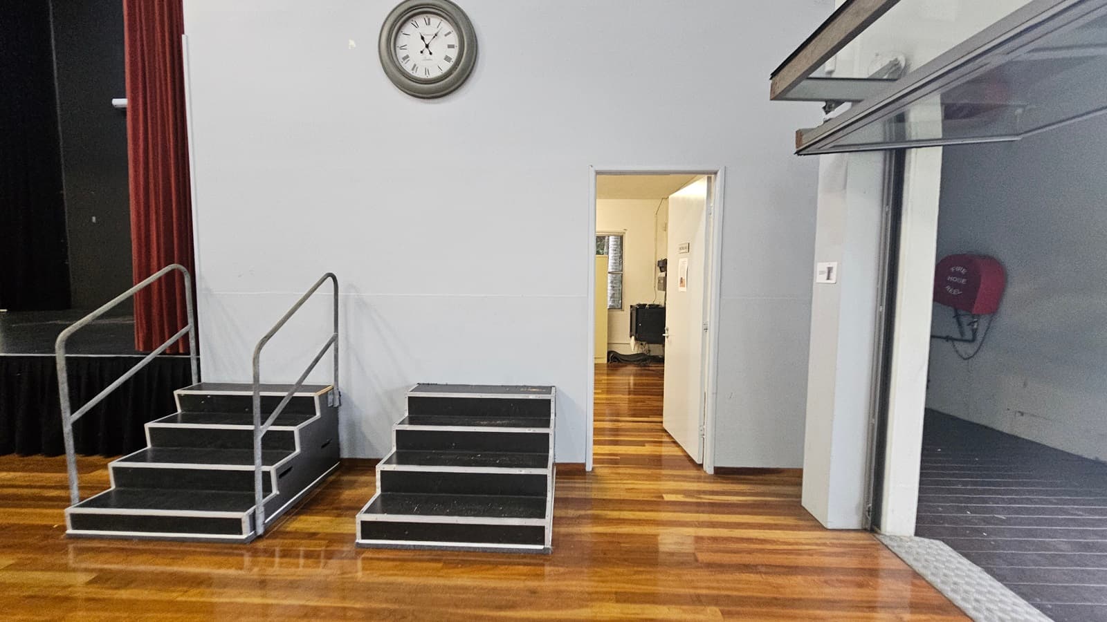 On the left, there are two sets of black steps with metal railings leading up to a stage, framed by red curtains. In the middle, a white door is open, revealing a room with wooden flooring and a window. A large round clock is mounted above the door. On the right, there's a grey wall with a red fire hose reel attached. The floor is polished wood, and the ceiling is not visible.